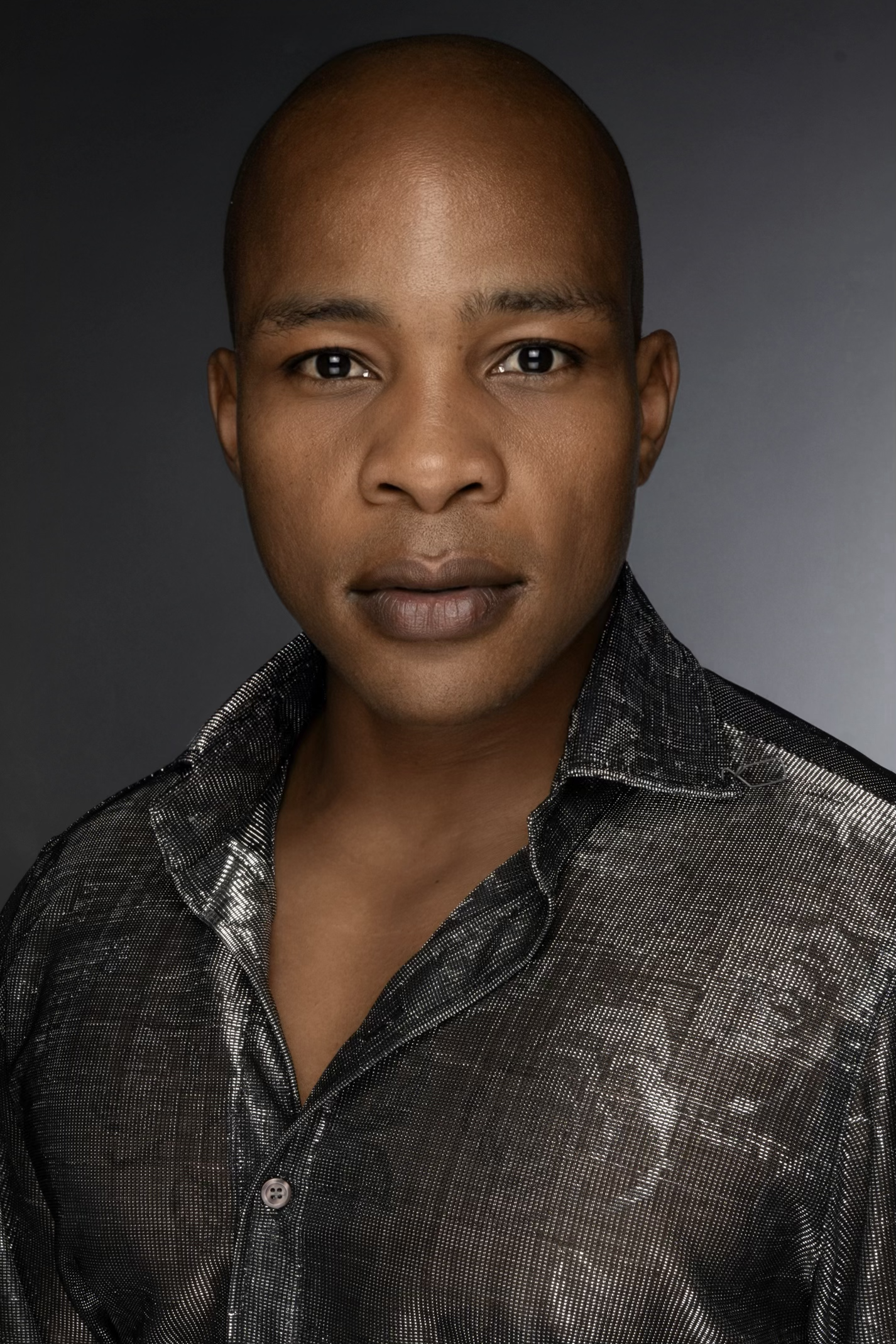 Close-up portrait of a woman with a shaved head, wearing a black and white patterned button-up shirt, against a dark background.