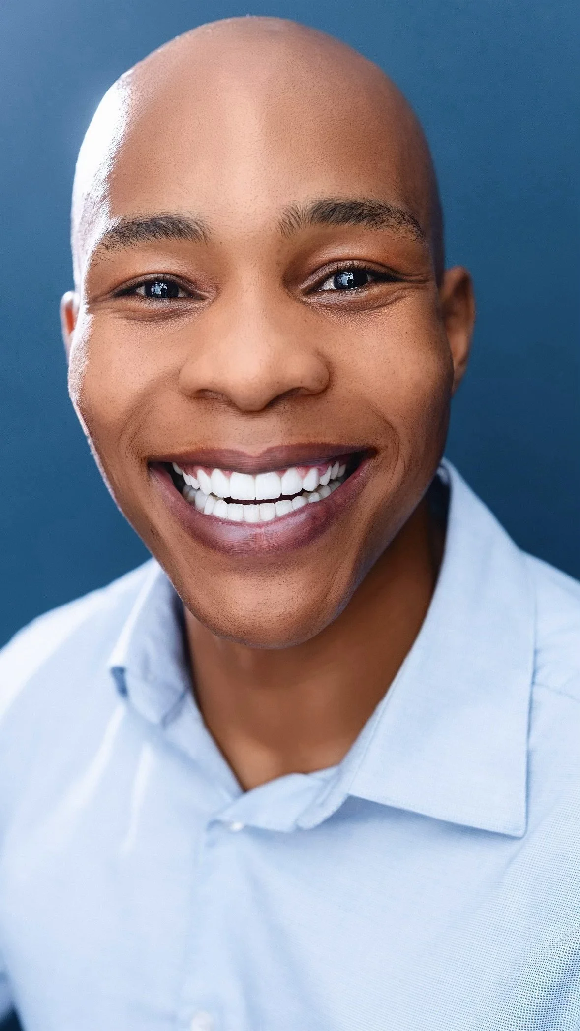 Close-up of a smiling person with a bald head, wearing a light blue collared shirt, against a blue background.