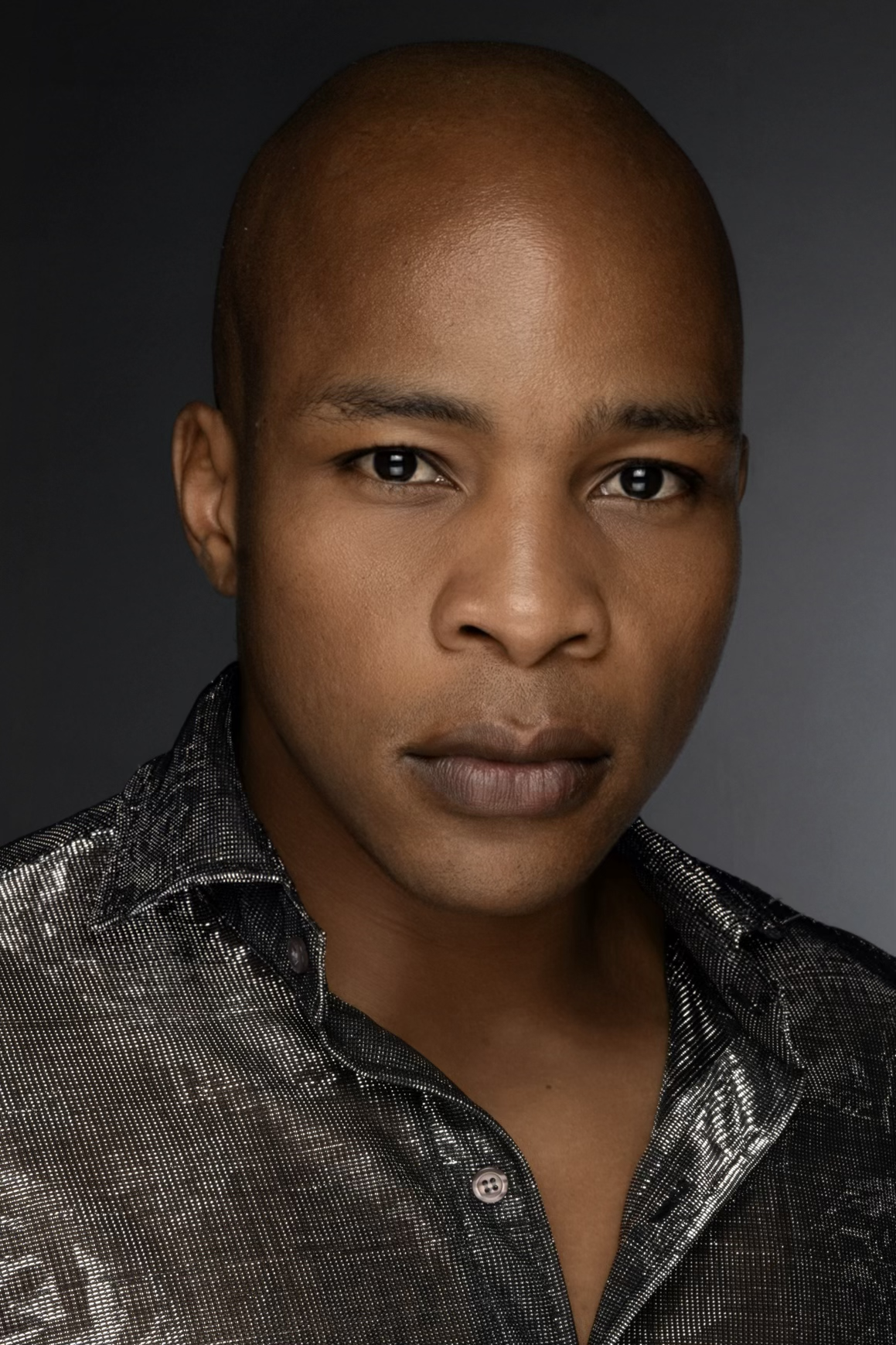 Close-up portrait of a man with smooth shaved head, dark eyes, and a confident expression, wearing a shiny black button-up shirt against a dark background.