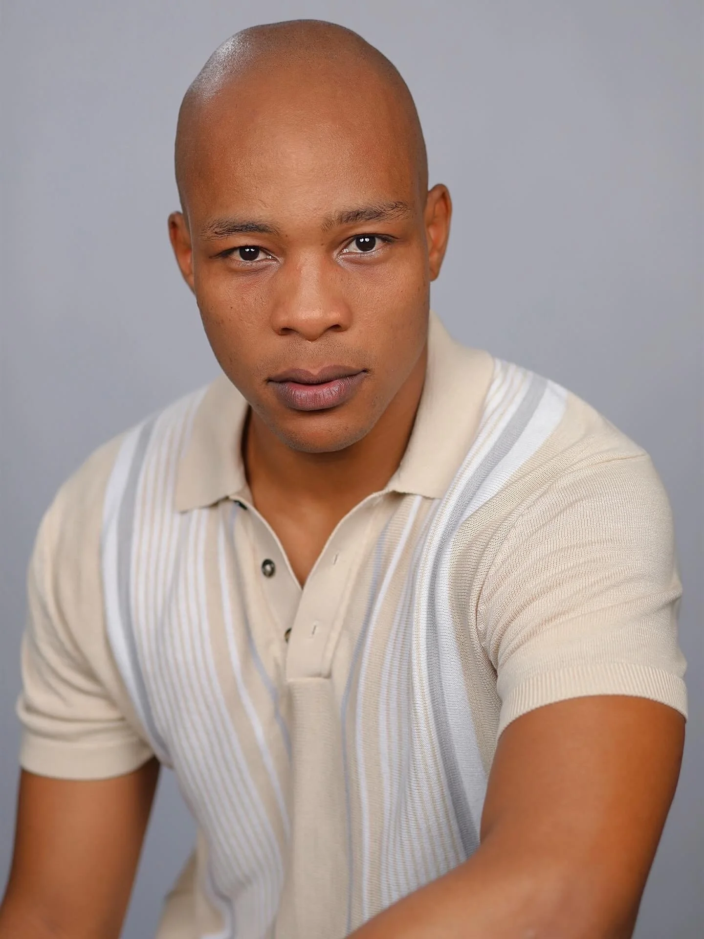 A young man with a shaved head wearing a cream and beige striped polo shirt, looking directly at the camera with a neutral expression, against a plain gray background.