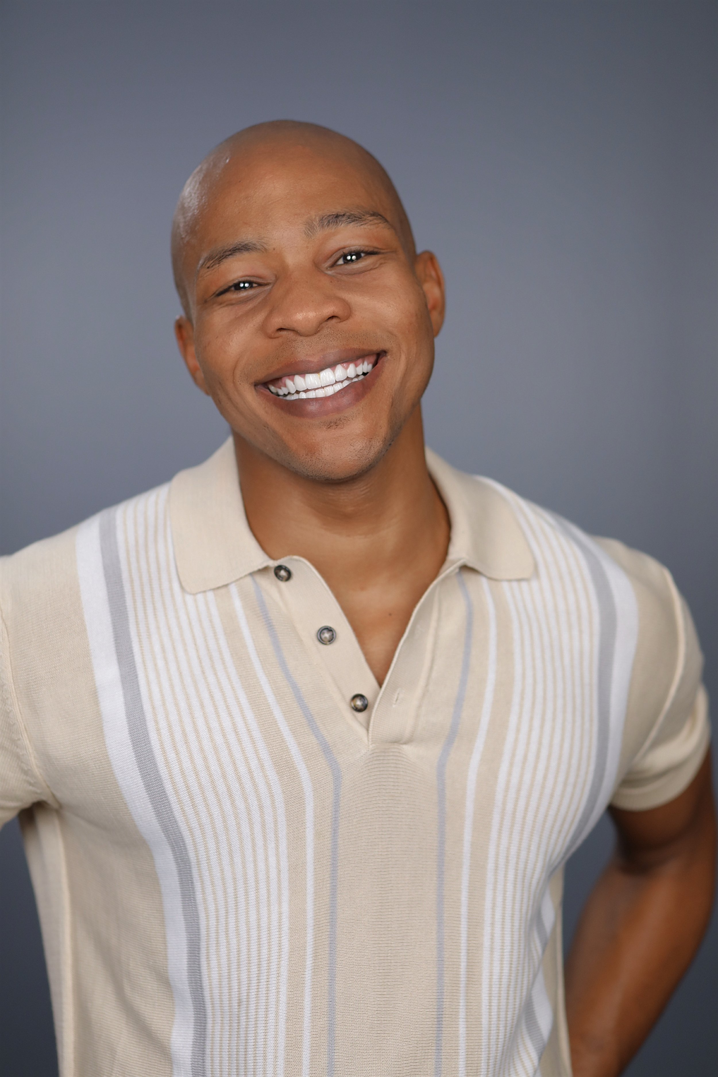 A smiling man with a shaved head wearing a beige polo shirt with vertical stripes, standing against a plain gray background.