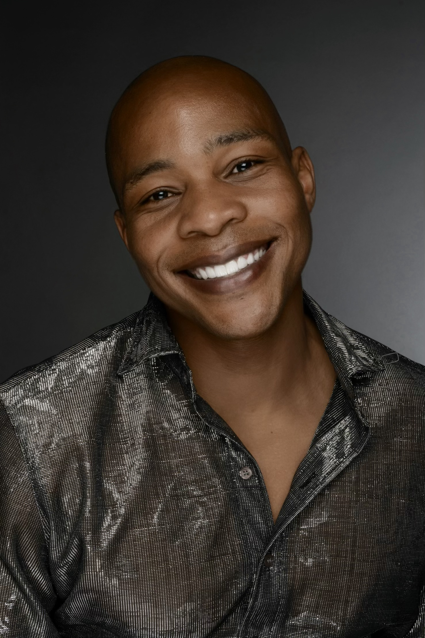 A person with a shaved head smiling, wearing a shiny, metallic patterned shirt, standing against a dark gray background.