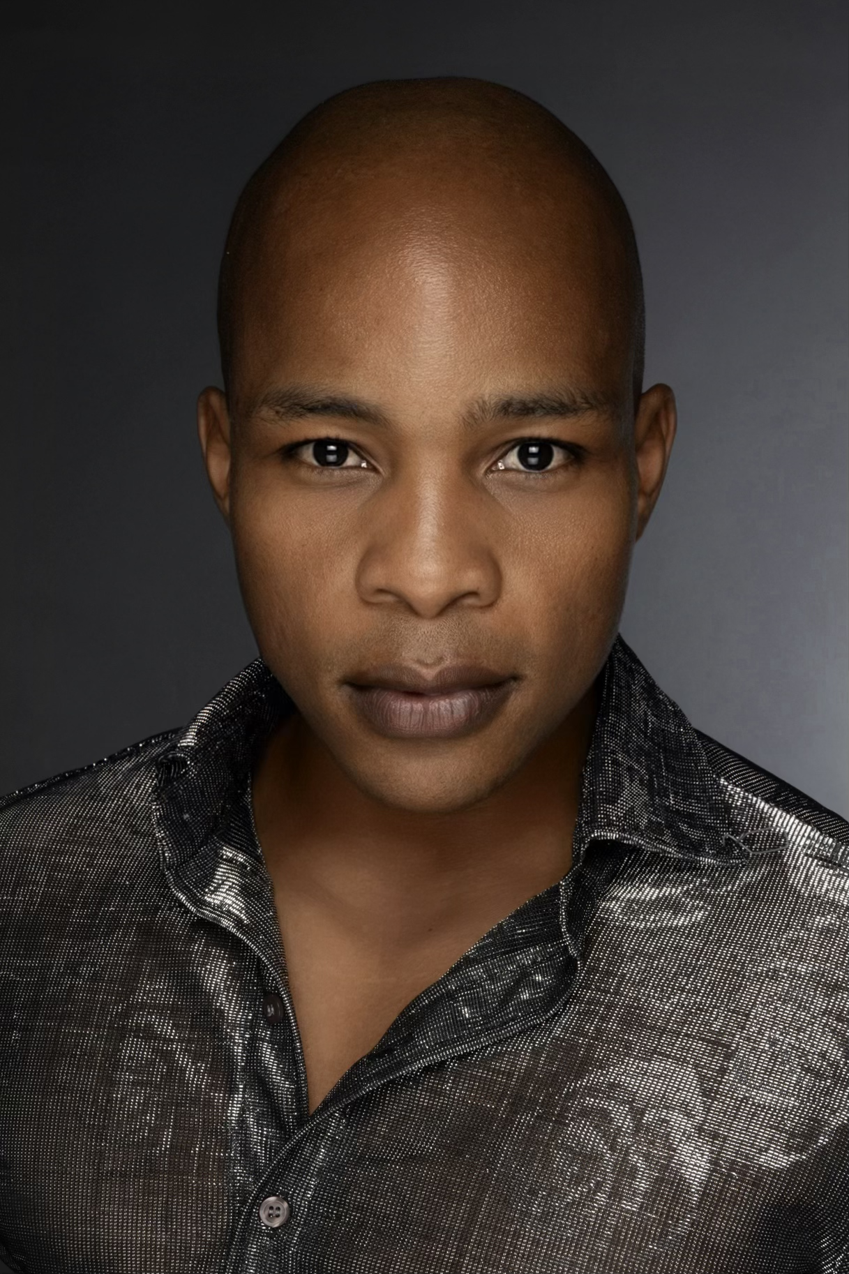 A close-up portrait of a bald man with medium brown skin, wearing a metallic black shirt with a shiny pattern, against a dark gradient background.