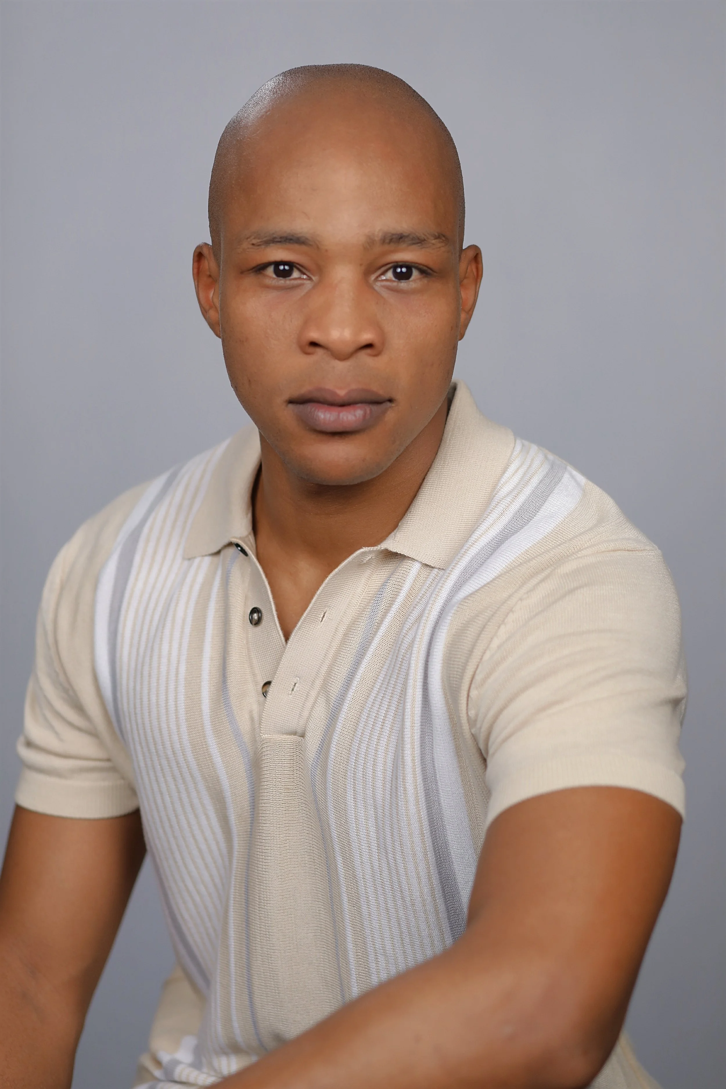 Close-up portrait of a young man with a shaved head, wearing a beige polo shirt with vertical stripes, against a plain gray background.
