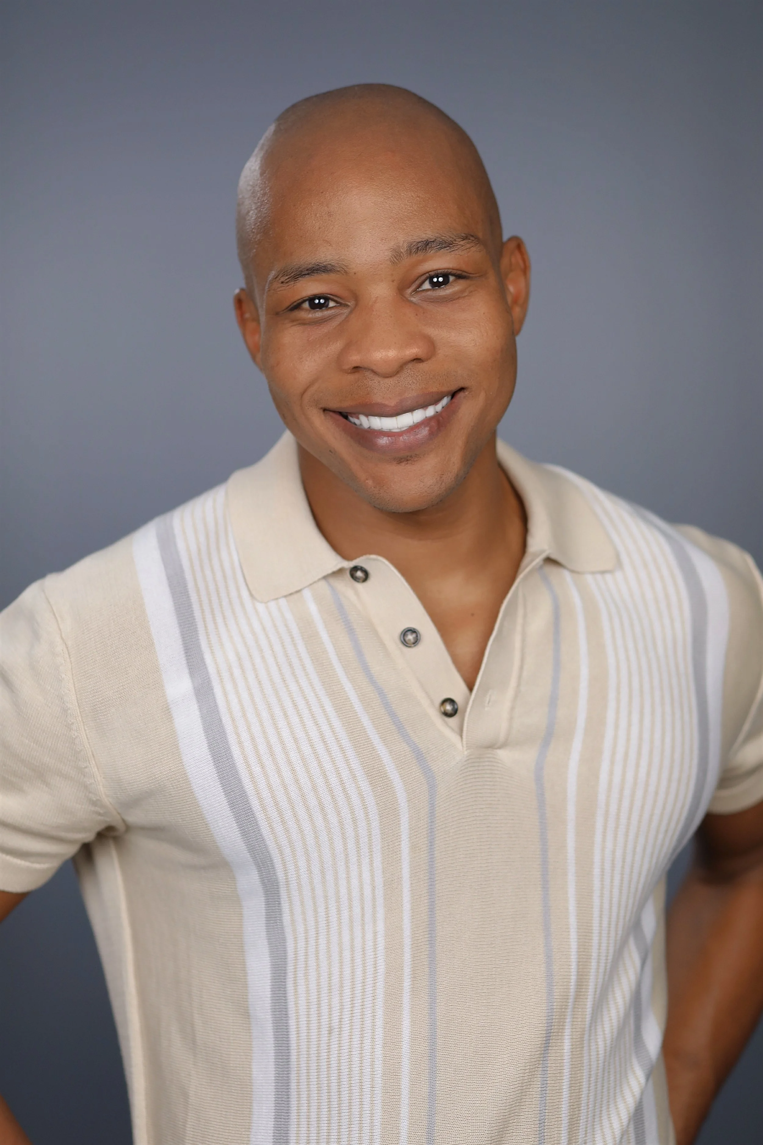 A smiling man with a shaved head and wearing a beige and white striped polo shirt, standing against a plain gray background.