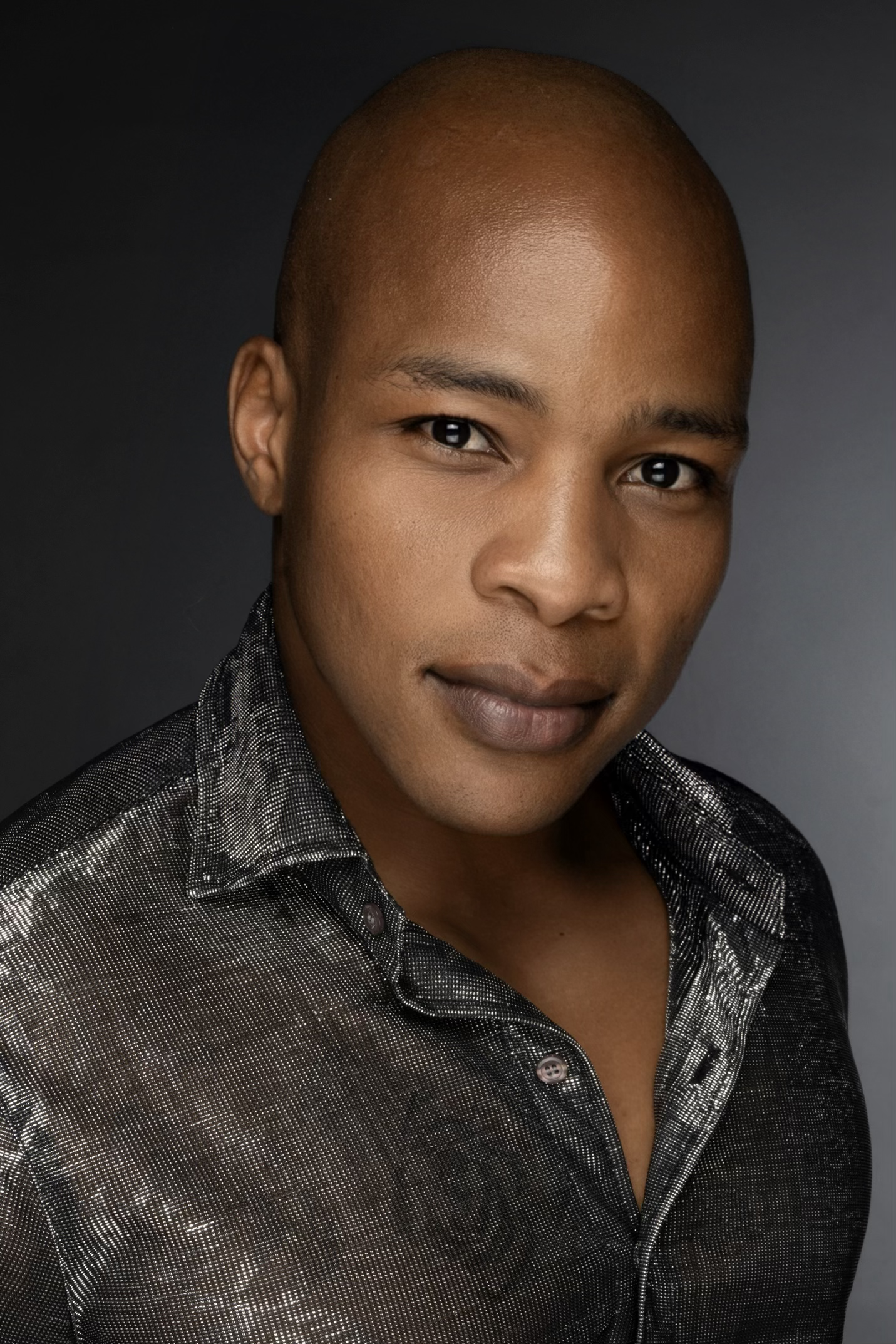 Close-up portrait of a person with a shaved head and dark skin, wearing a metallic-patterned shirt against a dark background.