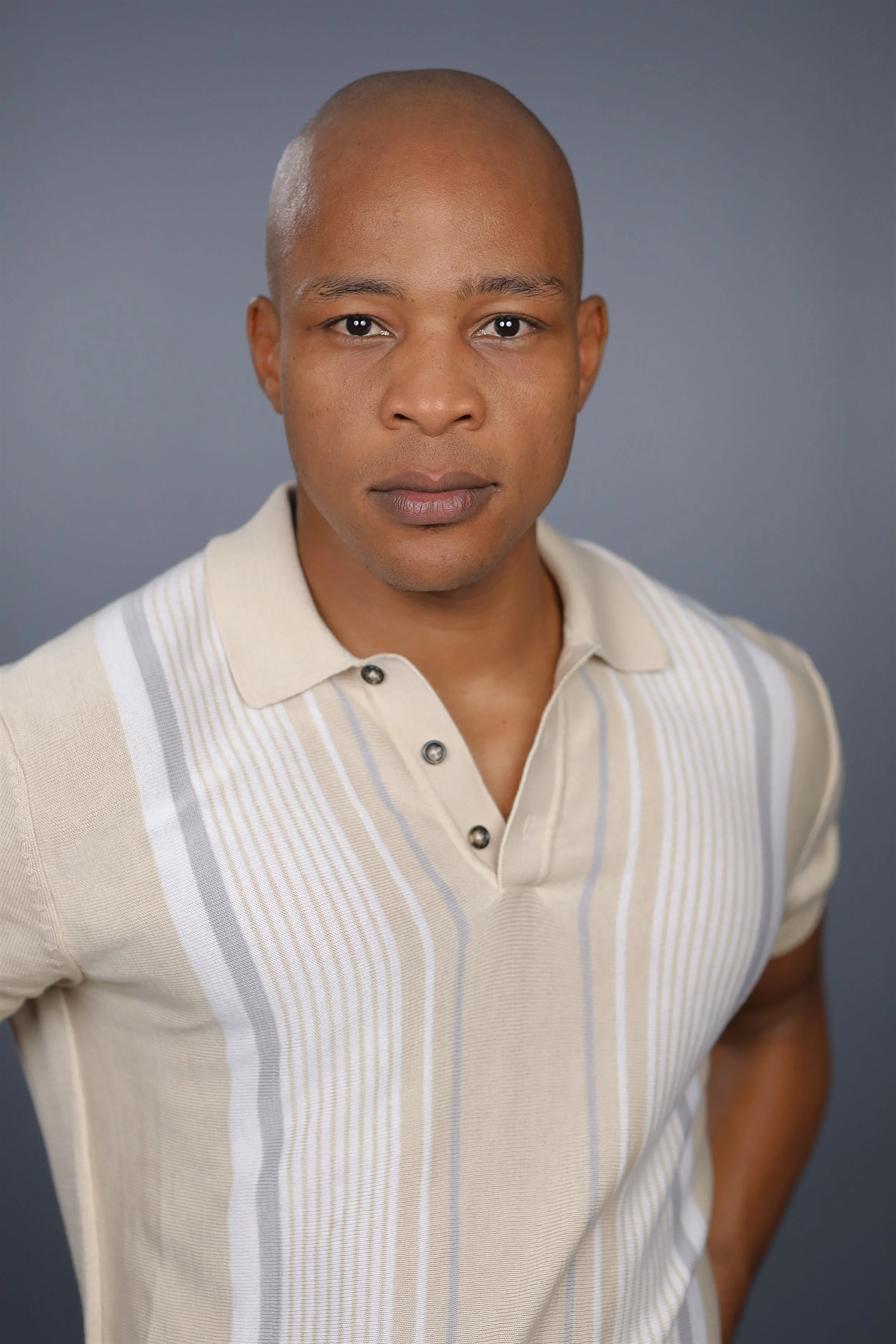 A man with a shaved head wearing a beige, striped polo shirt against a plain gray background.