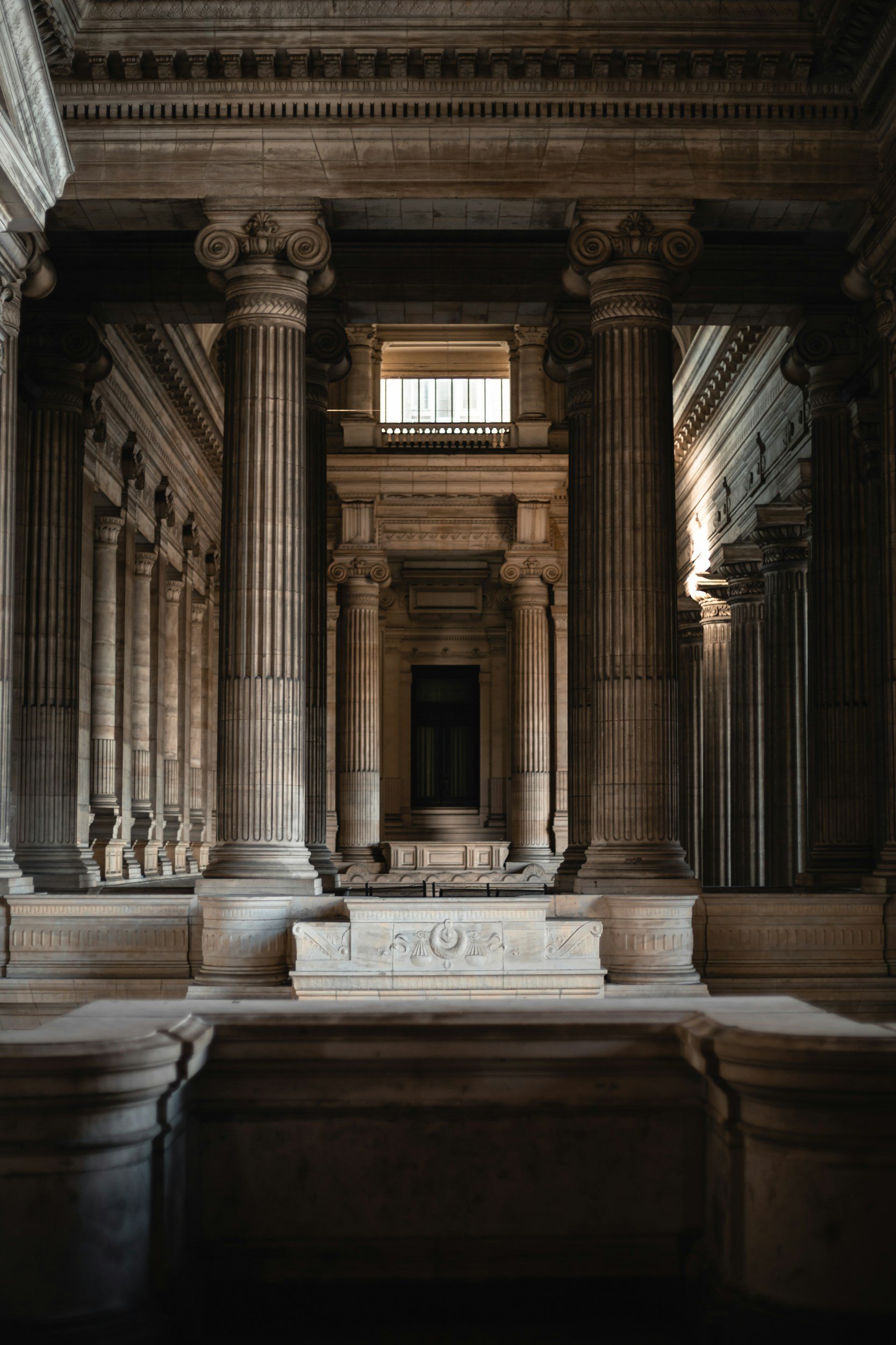 Interior of a grand classical building with tall marble columns, ornate architectural details, and a large window allowing natural light to illuminate the space.