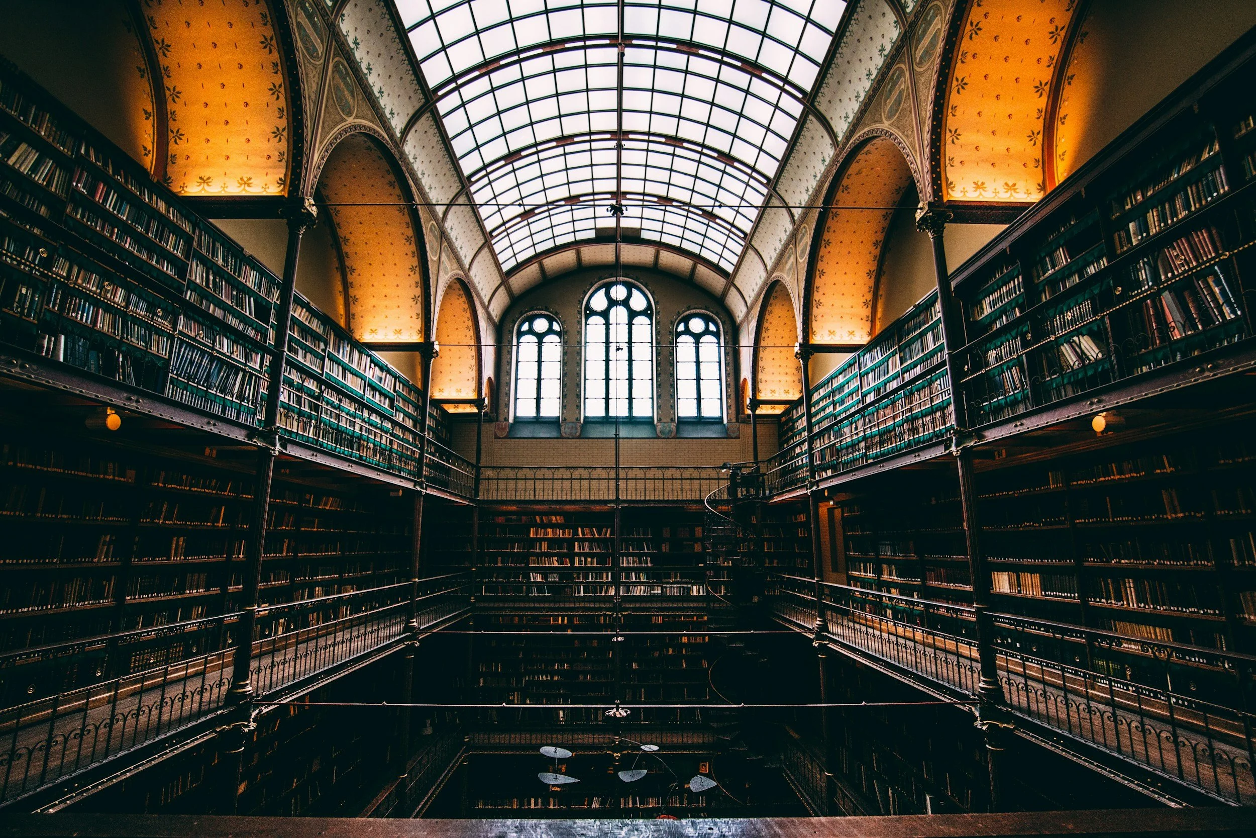Interior of a historic library with tall bookshelves filled with books, ornate yellow and gold arched walls, and a large glass domed ceiling letting in natural light.
