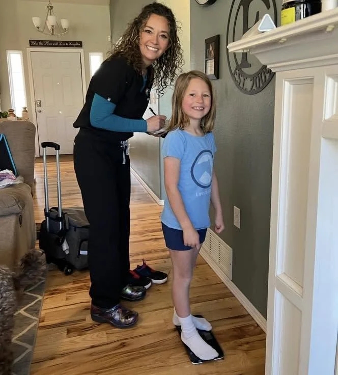 A woman with curly hair and a big smile stands next to a young girl. They are in a hallway of a home, with the woman appearing to sign something for the girl. The girl is smiling, wearing a blue T-shirt, shorts, and socks, standing on a small black and white scale. There is a suitcase and shoes beside them, and the home has hardwood floors, a beige couch, and wall decorations.