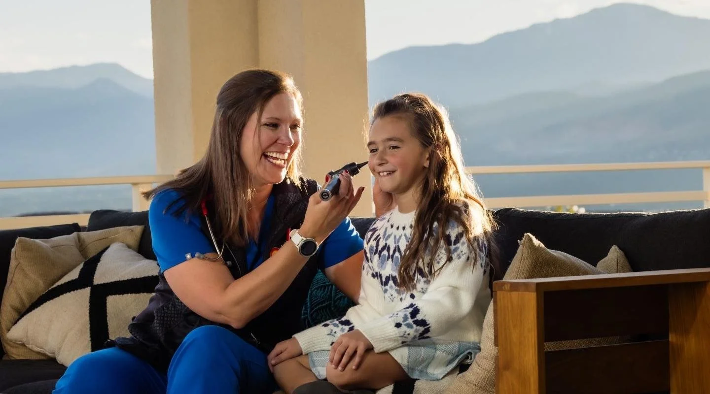 A healthcare professional, possibly a nurse or doctor, examines a young girl with an otoscope while seated on a couch. The girl is smiling and appears comfortable. The setting is a balcony or porch with mountain scenery in the background.