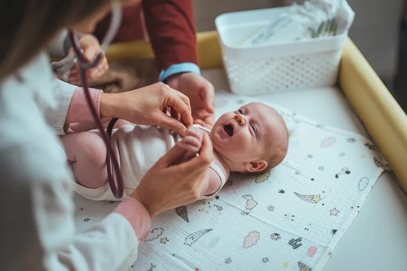 A baby lying on a changing table receiving a checkup from a healthcare professional, with a stethoscope placed on the baby's chest.