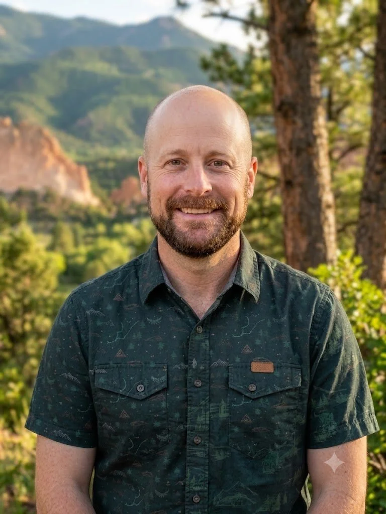 A smiling man with a beard and bald head standing outdoors in a forested area with mountains in the background.