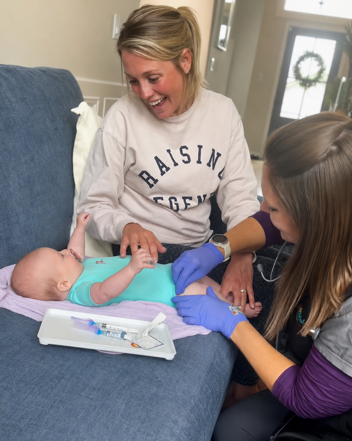 A woman and a nurse are tending to a baby lying on a couch. The woman is smiling and the nurse is administering a vaccination with a syringe. The baby is lying on a purple towel with medical supplies nearby.