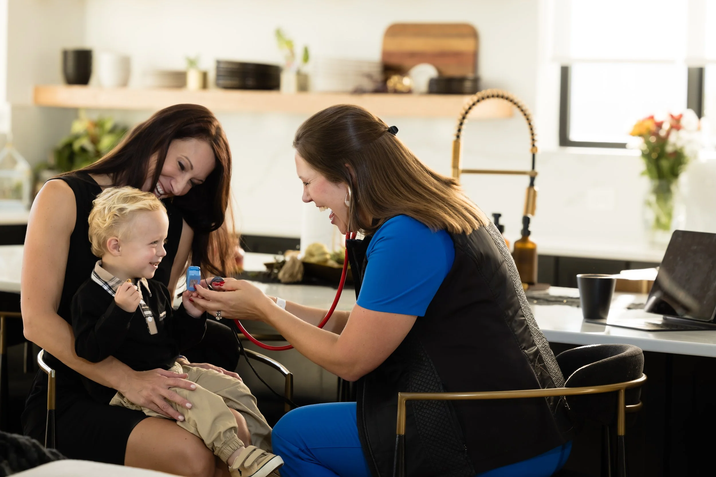A healthcare professional using a stethoscope to examine a young boy, with a woman and another woman smiling nearby in a bright, modern kitchen.