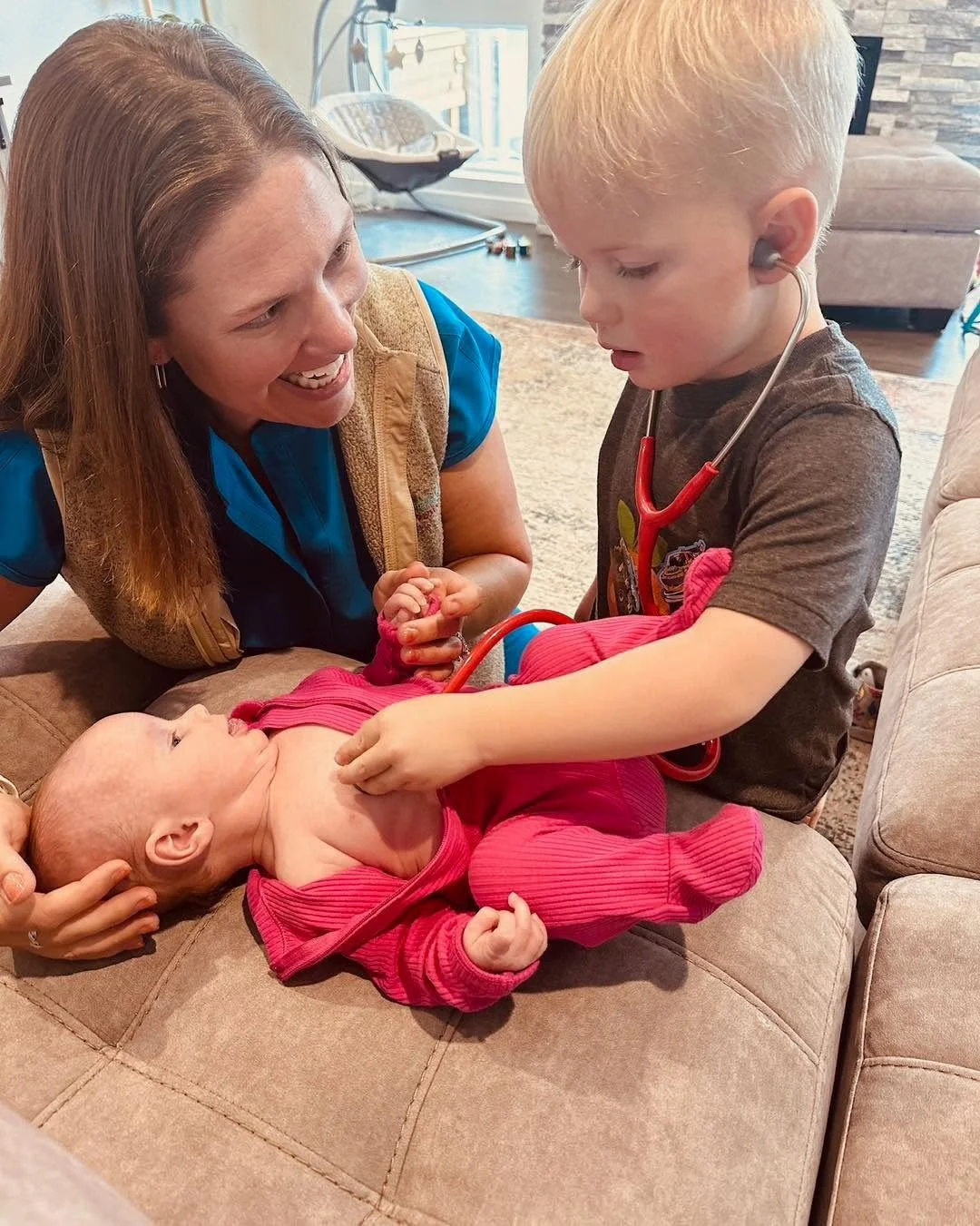 A woman, a young boy with a stethoscope, and a baby lying on a couch. The boy appears to be playing doctor, listening to the baby's chest. The woman is smiling and watching them.