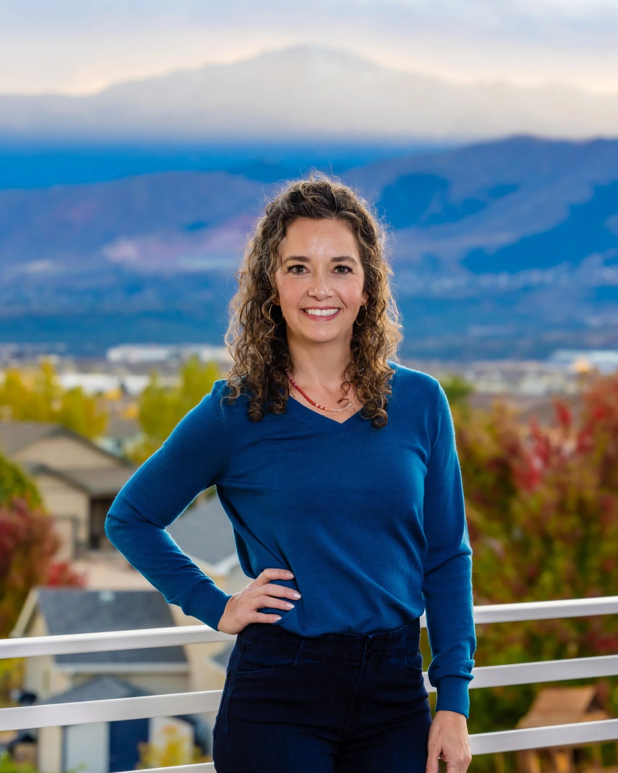 A woman with curly brown hair smiling and standing outdoors, wearing a blue top and black pants, with mountains and autumn trees in the background.