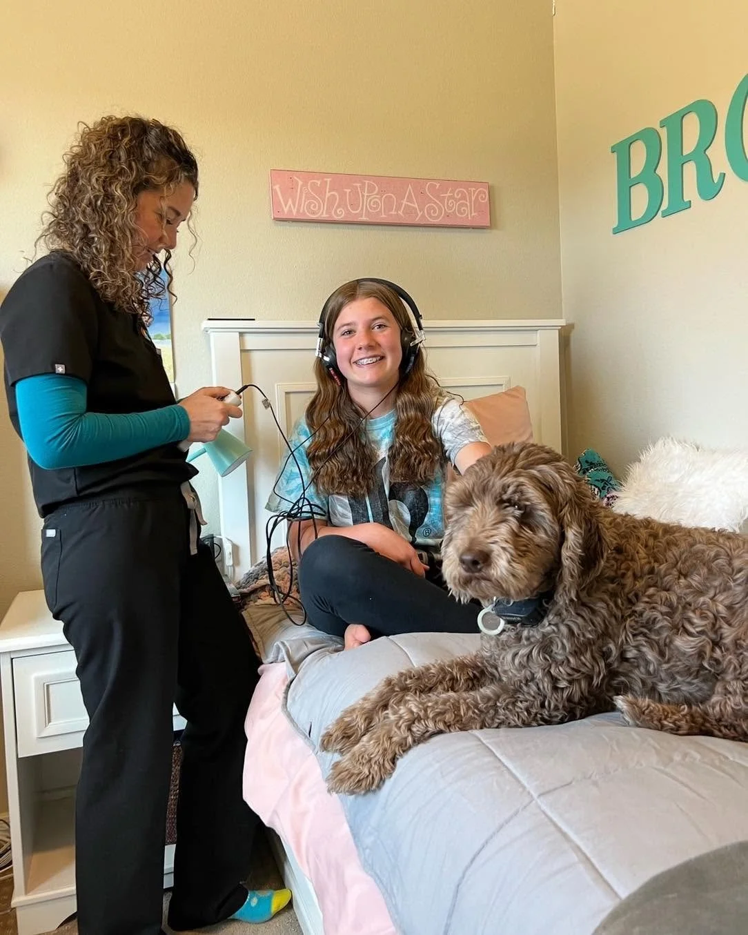 A girl with a headset sitting on a bed, smiling, with a large brown dog lying next to her. A woman in scrubs is standing nearby, holding a medical device.
