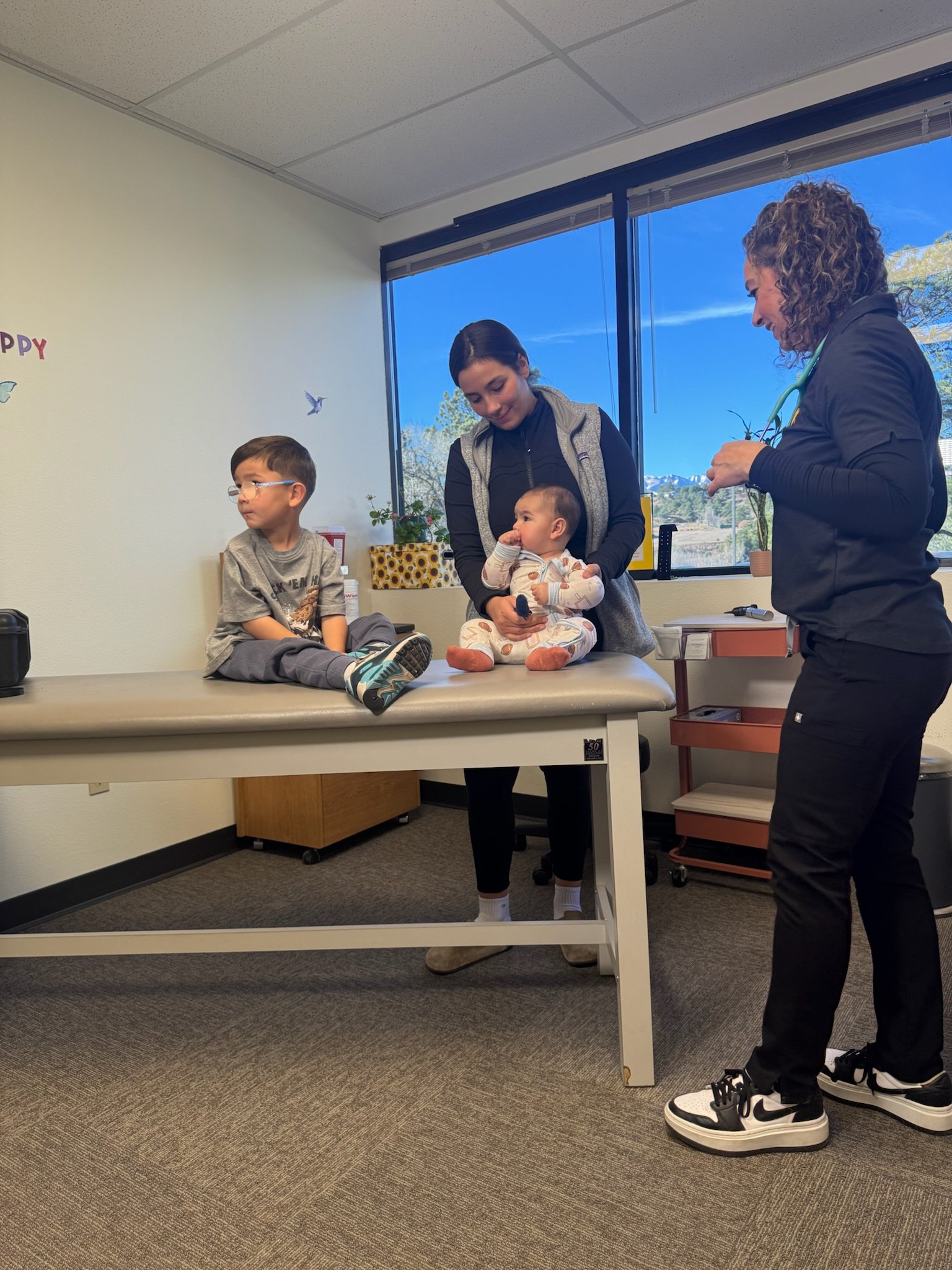 A doctor examining a baby on an examination table with a woman holding the baby. A young boy is sitting on the table nearby, looking away. The scene takes place in a medical office with a large window showing a view of trees and mountains in the background.
