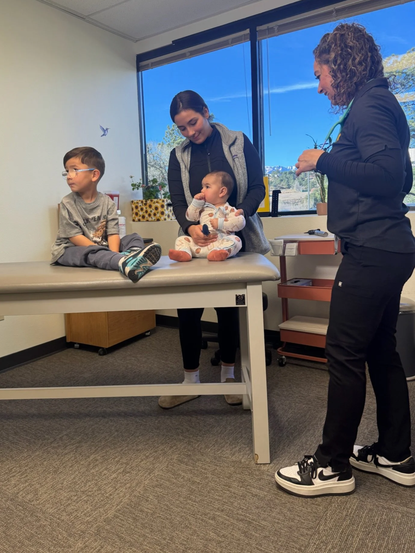A young woman is holding a baby boy on an examination table in a clinic or hospital room. A boy with glasses is sitting on the edge of the table, and a nurse or doctor is standing nearby. The room has a window showing a clear sky and mountains outside, with posters, plants, and medical equipment visible.