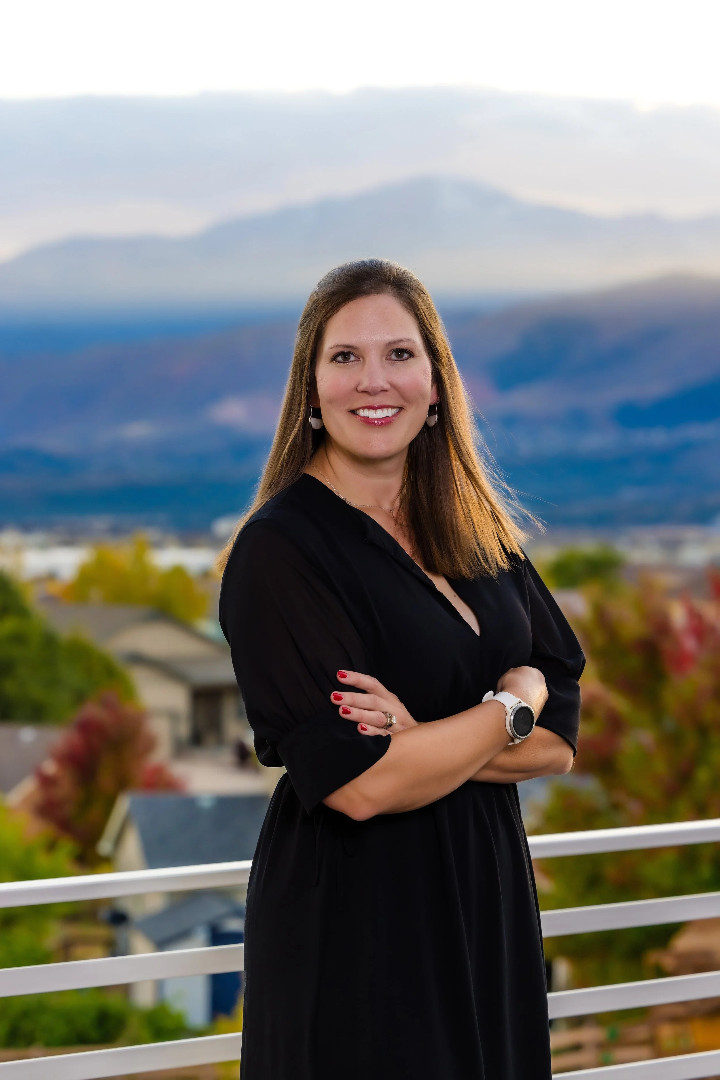 A smiling woman with long, brown hair, wearing a black dress, earrings, a watch, and a ring, standing with arms crossed in front of a mountainous landscape with a cloudy sky.