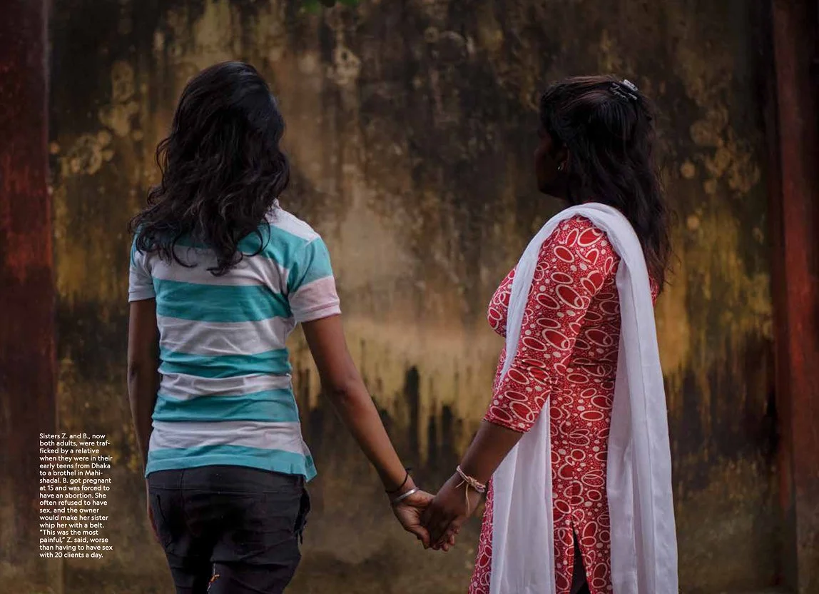 Two women holding hands, one in a striped shirt and the other in a patterned dress with a white dupatta, standing against a textured wall.