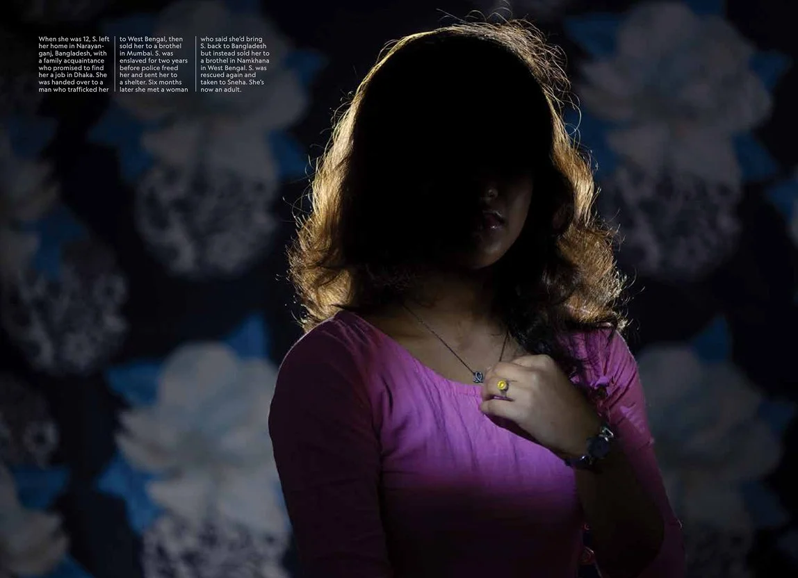 A young woman with wavy hair stands in front of a backdrop decorated with large, dark and white floral patterns, partially obscuring her face with shadows, as she looks downward, wearing a purple top.