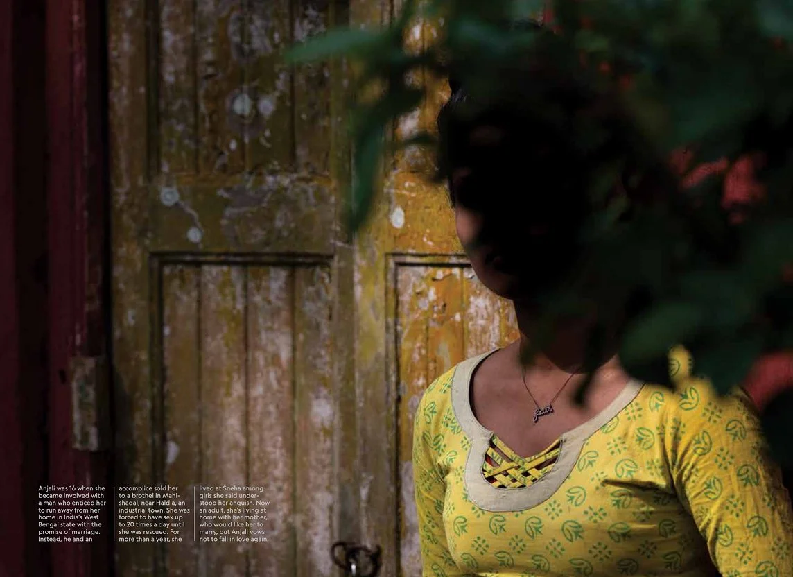 A young woman in a yellow shirt with green pattern standing in front of a rustic wooden door, partially obscured by green leaves in the foreground.