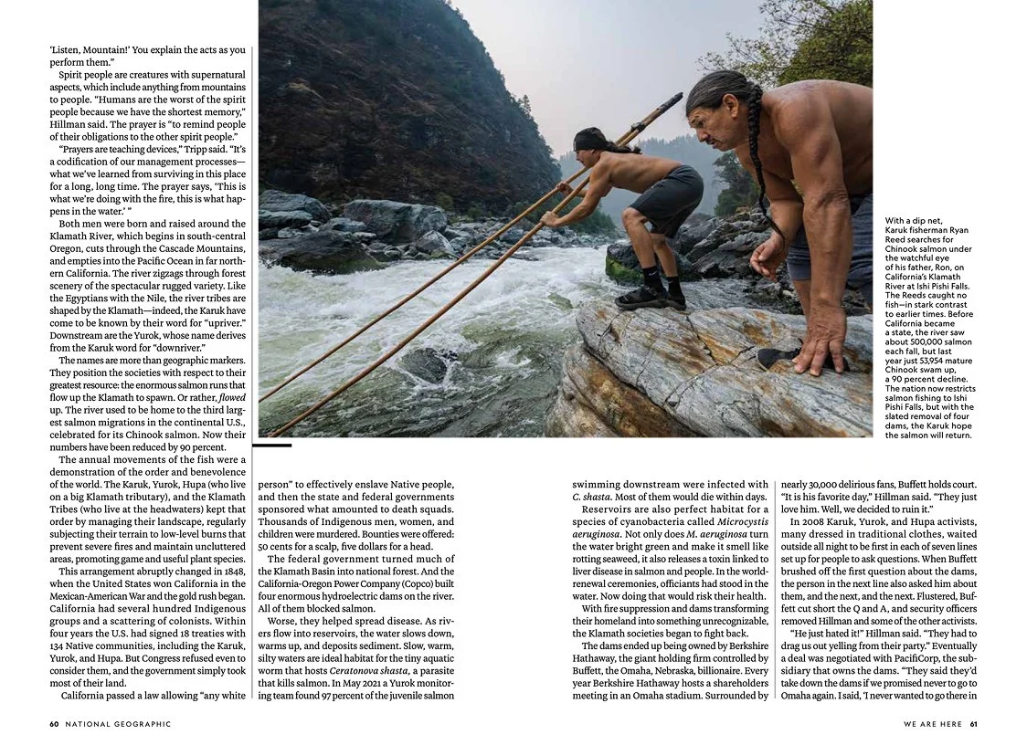 Two people, a man and a woman, are hot on the trail of a river and surrounded by steep, forested mountains in the background. The woman is in front, crouched over a large rock, holding a stick, composed and ready for action, while the man is behind, leaning forward, observing, with his hand on a rock. They are dressed in outdoor clothing suitable for hiking or exploring.