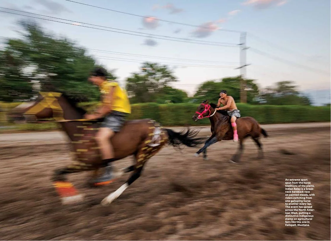 Two riders racing on a dirt track; one rider is shirtless with pink leg wraps on a brown horse, the other rider in a yellow shirt on a galloping horse; motion blur indicates high speed; background has power lines and trees.