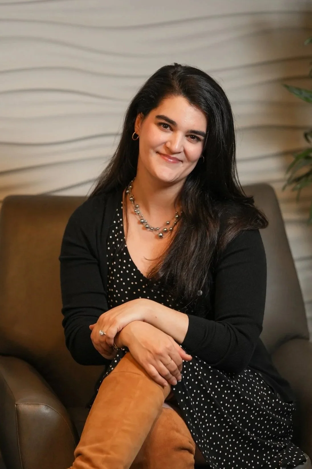 A woman with long dark hair, wearing a black and white polka dot dress and a black cardigan, sitting on a brown couch with a textured beige wall and green plant in the background.