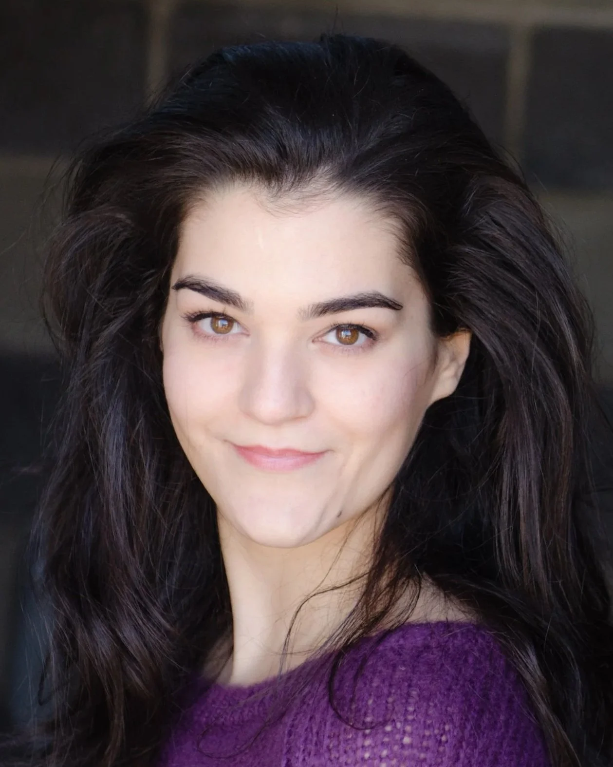 A young woman (actor Lauren Ezzo) with long dark hair and light skin, smiling softly, wearing a purple sweater.