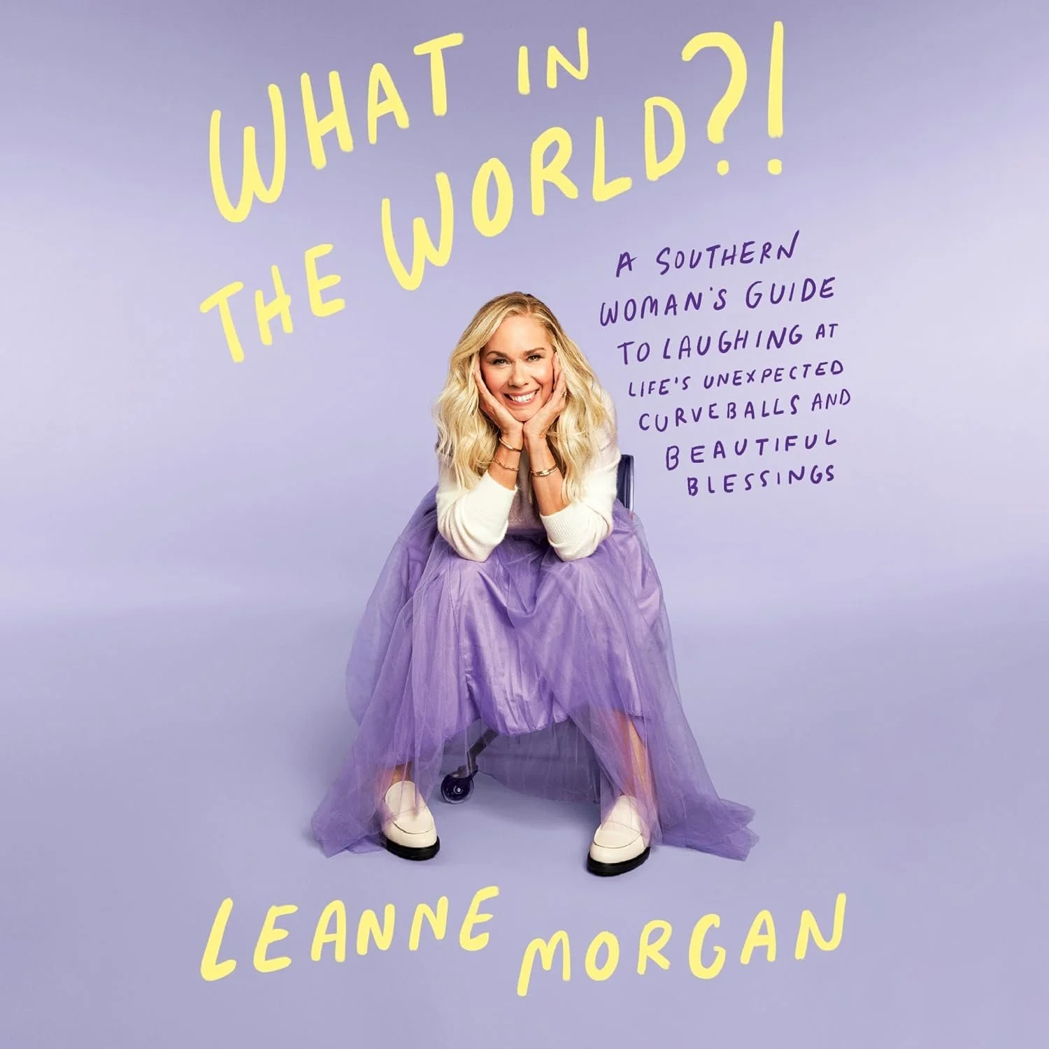Book cover featuring a woman with long blonde hair smiling, sitting on a chair with purple tulle skirt, against a purple background. Title: "What in the World?!" in yellow text and subtitle: "A Southern Woman's Guide to Laughing at Life's Unexpected Curveballs and Beautiful Blessings" in purple. Author: Leanne Morgan in yellow text.