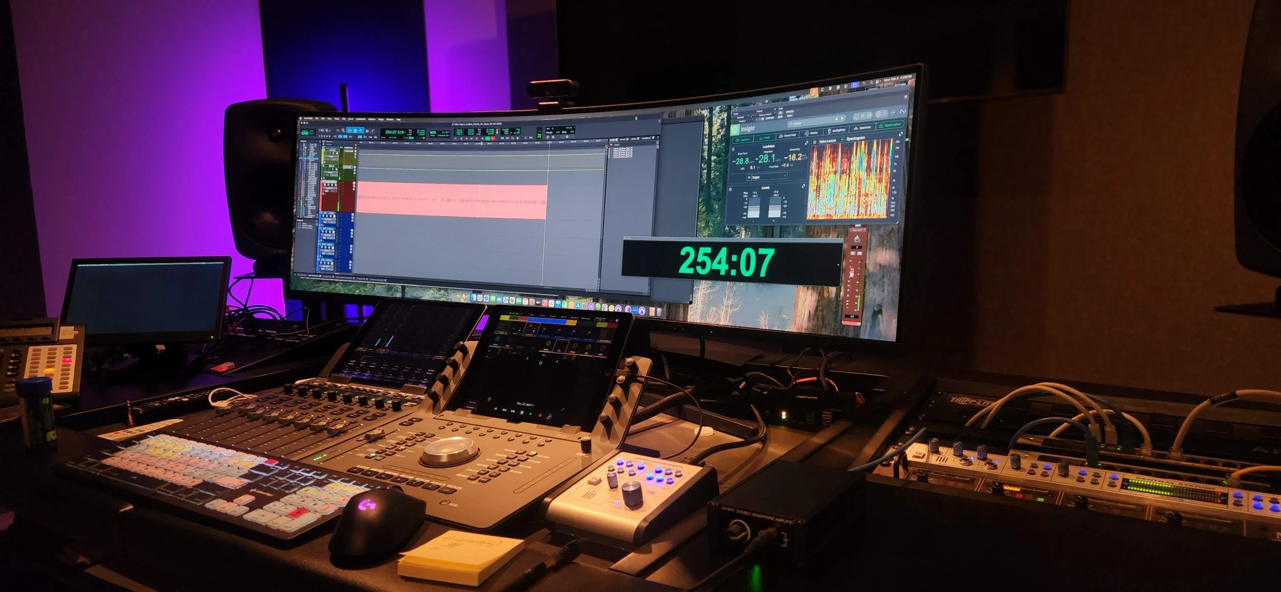A music production studio mixing console with multiple screens displaying audio editing software, surrounded by audio equipment, in a dimly lit room with purple and blue lighting.
