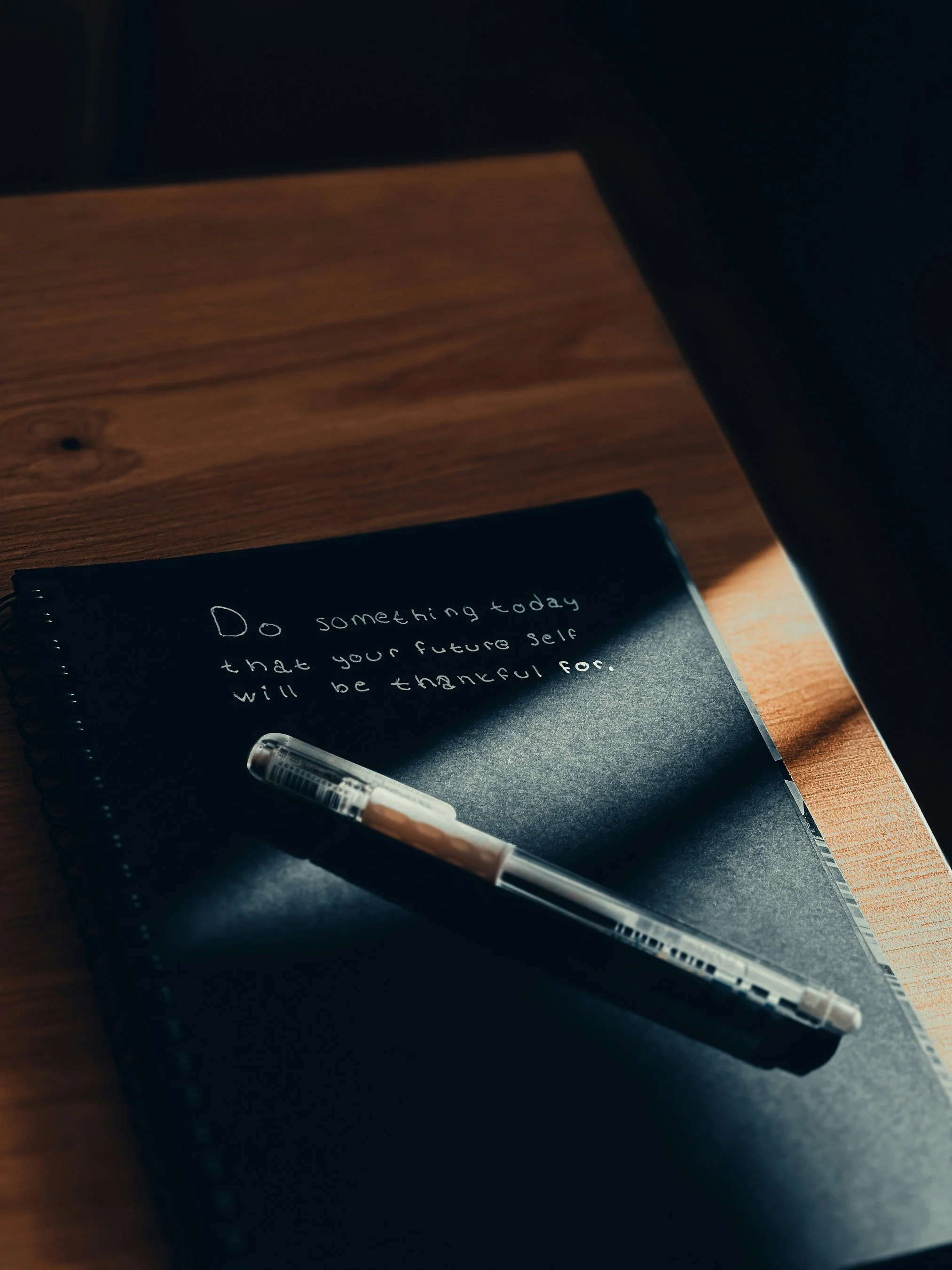 A black notebook with white handwritten text, a pen resting on top, placed on a wooden surface. The text on the notebook says, 'Do something today that your future self will be thankful for.'