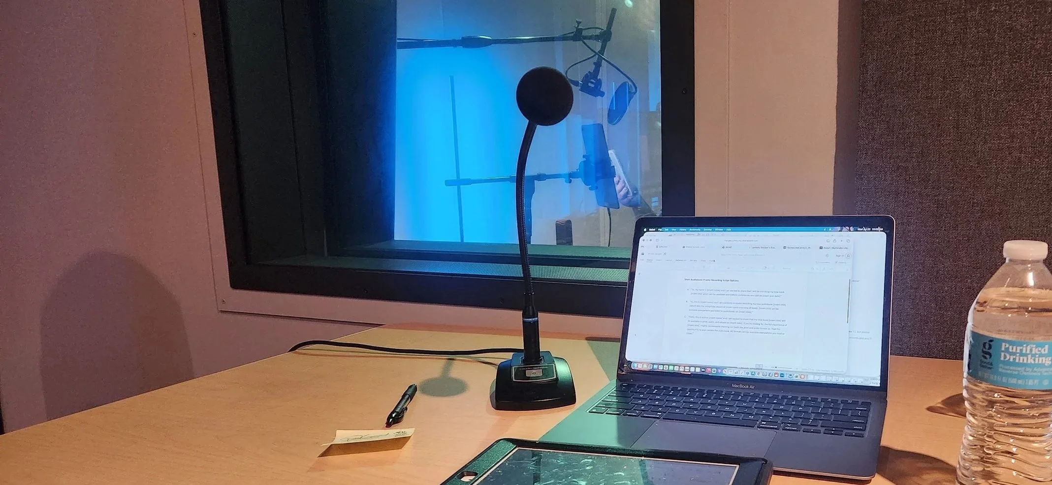 A recording studio observation room with a microphone, laptop, marker, and water bottle on the table. The room has a window looking into a recording booth with a microphone and stand visible.