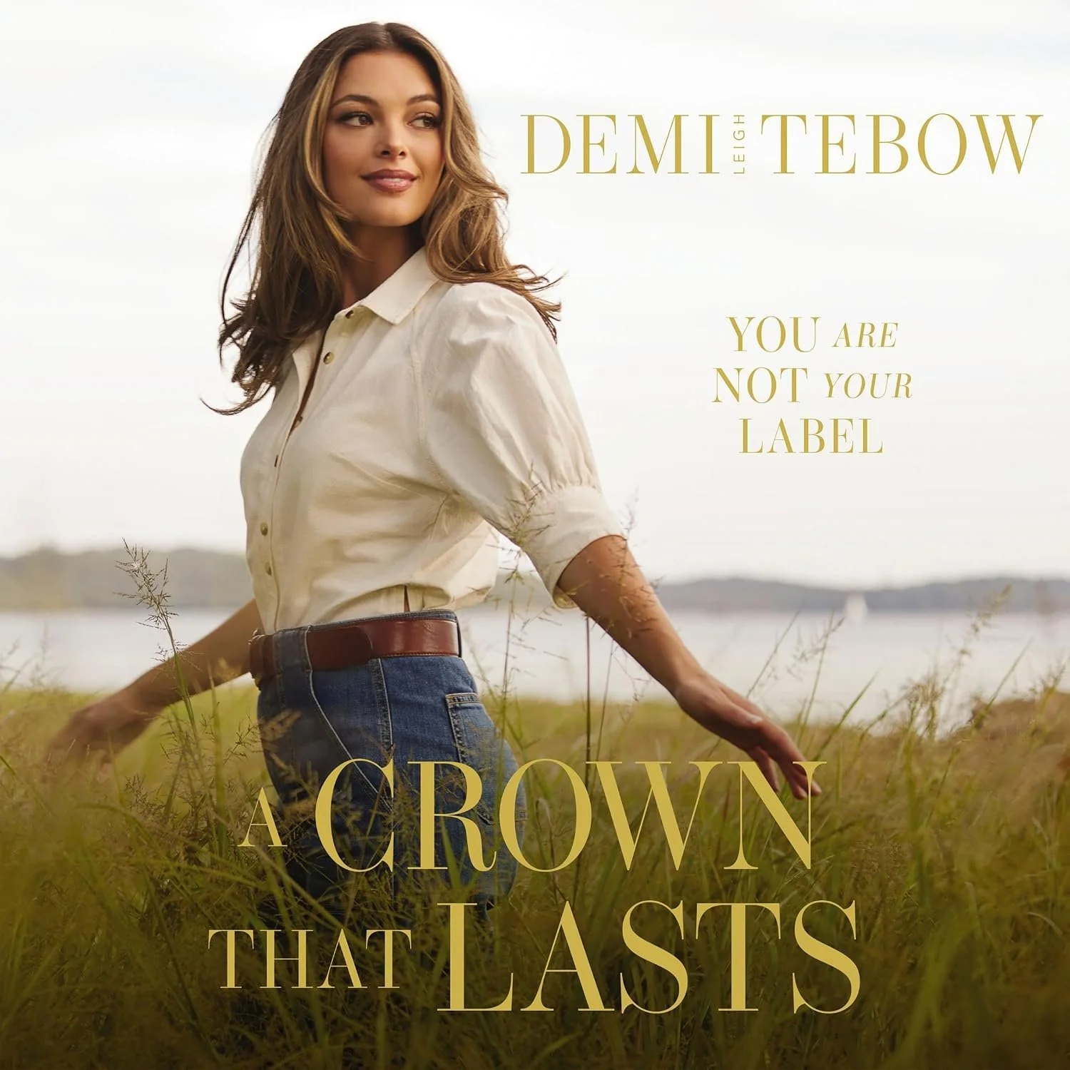A woman standing in a field of tall grass by a body of water, looking to the side, with text overlay that reads 'Demi Leigh Tebow - You are not your label. Grown that lasts.'