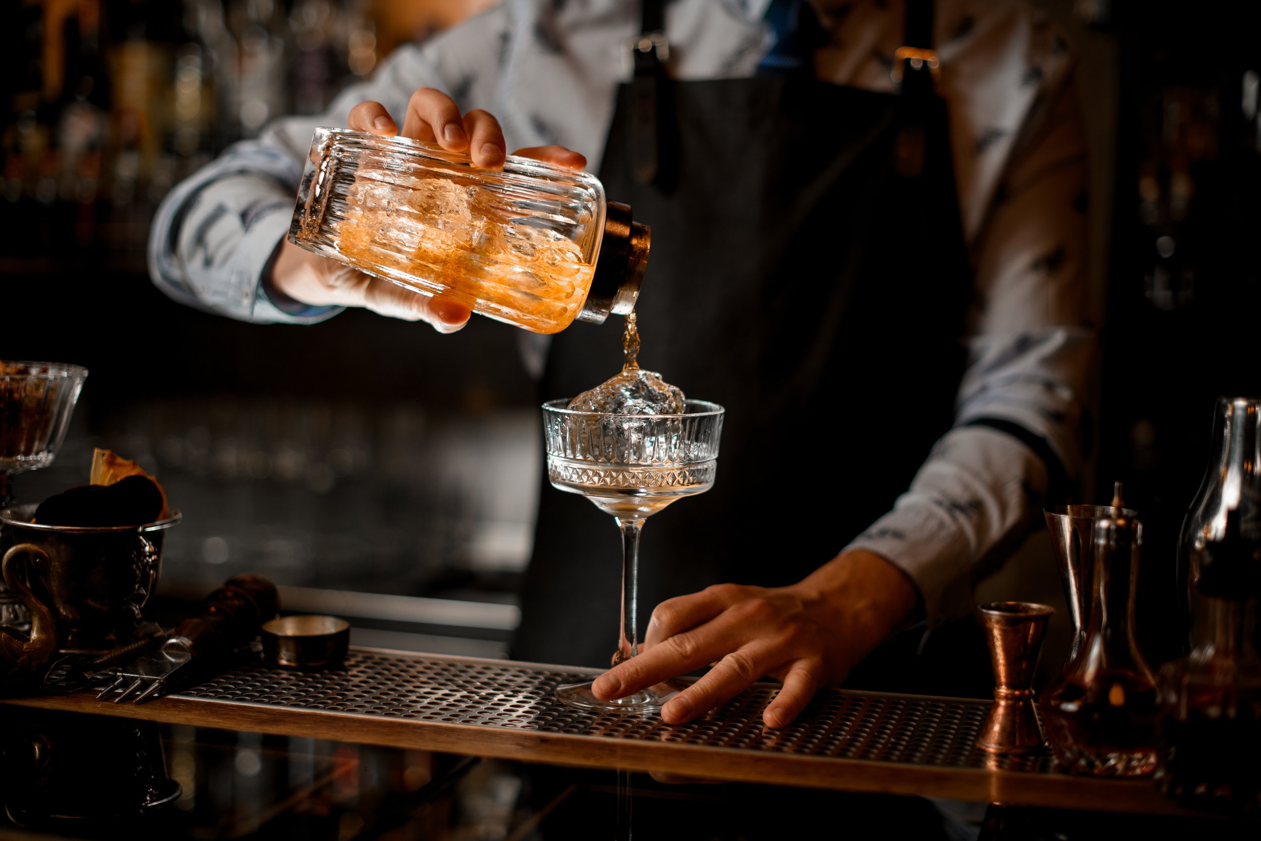 a bartender pictured from the shoulders down with a black apron and gray dress shirt pouring a drink at a bar