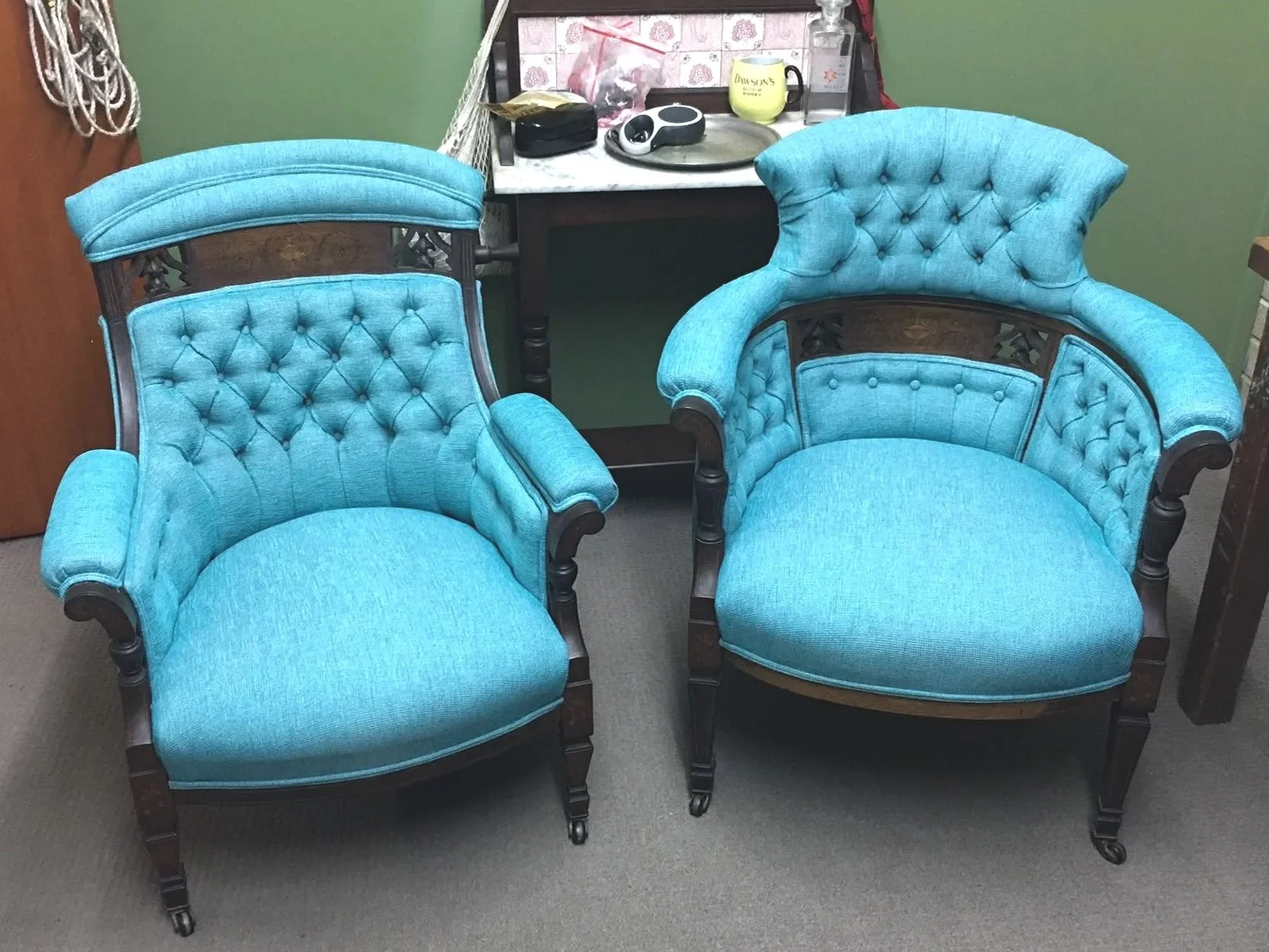 Two vintage teal armchairs with wooden frames and tufted upholstery sit side by side in a room. Behind them is a small black table holding various objects, including a cup, a remote, and small containers. The wall behind the table is partly covered w