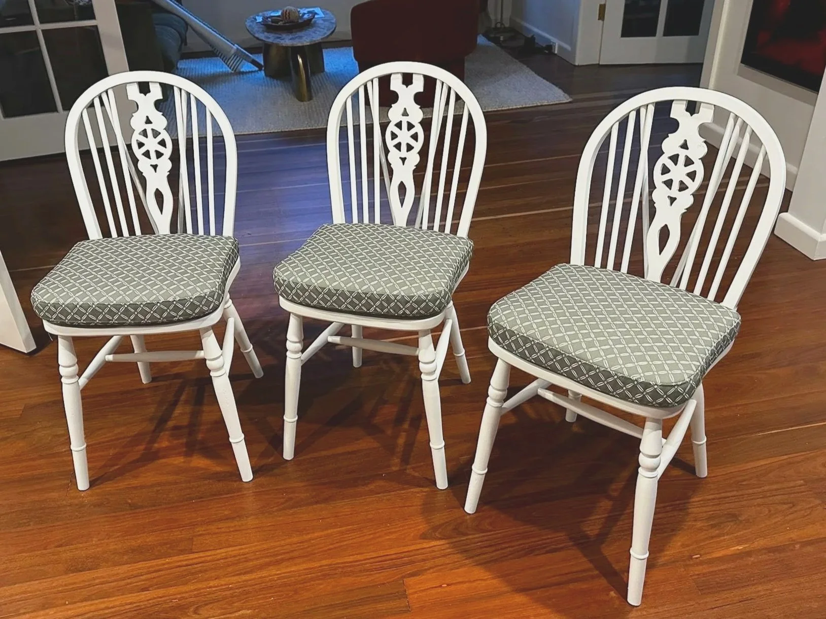 Three white wooden chairs with patterned cushions are arranged in a row on a hardwood floor in a dining room.