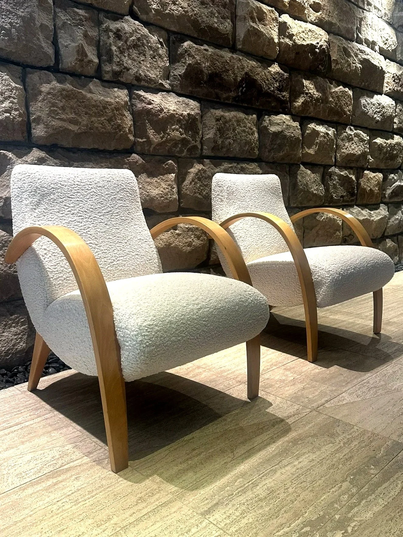 Two white upholstered chairs with wooden armrests and legs are positioned in front of a stone brick wall in a waiting area.
