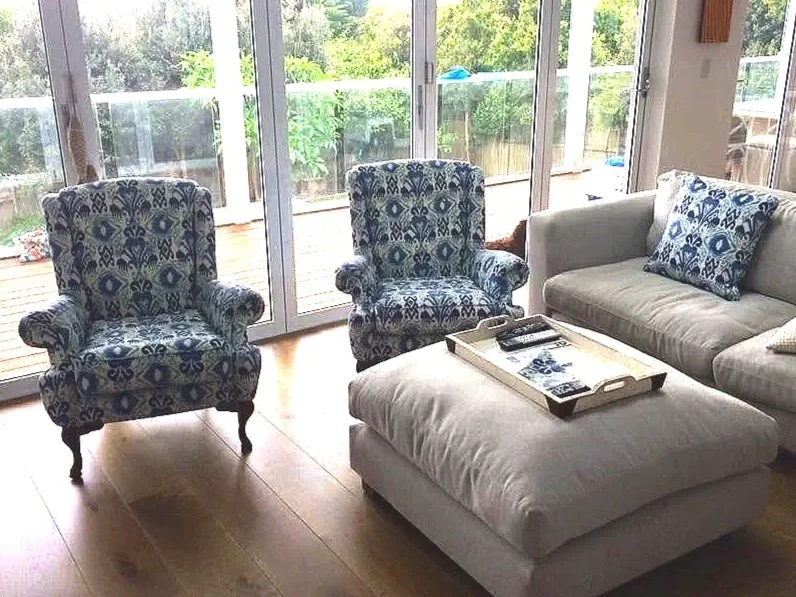 Living room with two patterned armchairs, a white sofa with a matching cushion, a white ottoman, tray with remote controls, and sliding glass doors leading to a patio with greenery outside.
