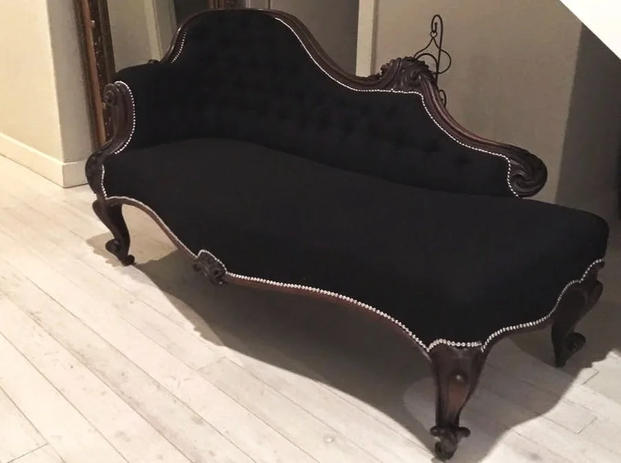 A vintage black velvet settee with ornate wooden carvings and white nailhead trim, positioned on a light-colored wooden floor.
