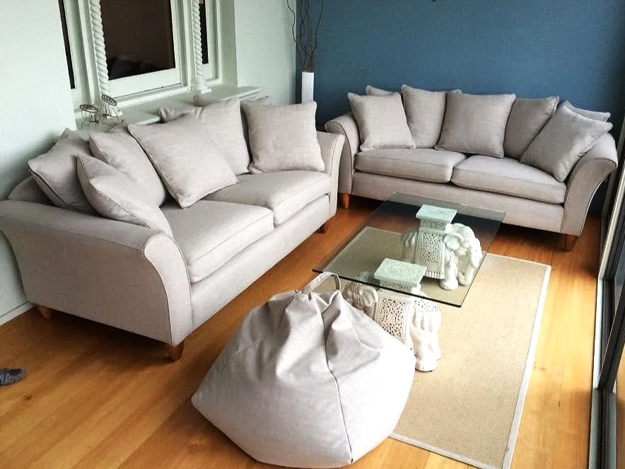 Living room with two beige sofas, a glass coffee table, a beige bean bag chair, decorative pillows, a cream-colored rug, a window with blinds, and a blue accent wall.