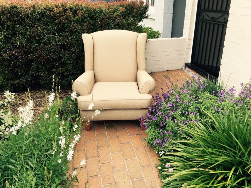 A beige armchair placed on a brick pathway next to purple and white flowering plants, with a hedge and white brick wall in the background.