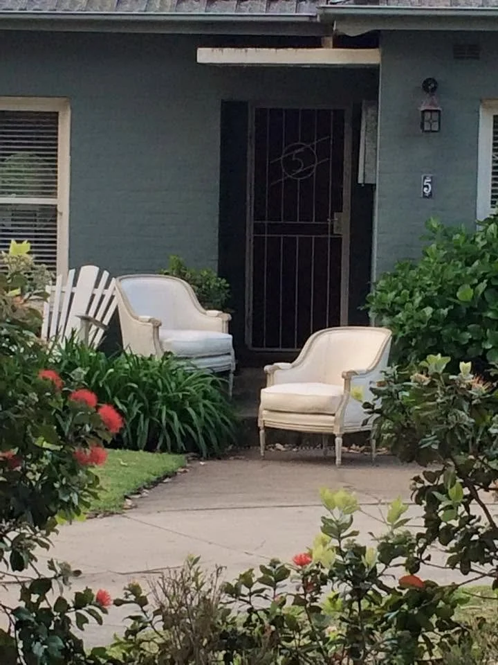 Front porch with two white vintage armchairs, surrounded by green bushes and flowering plants, with a gray house and black front door in the background.