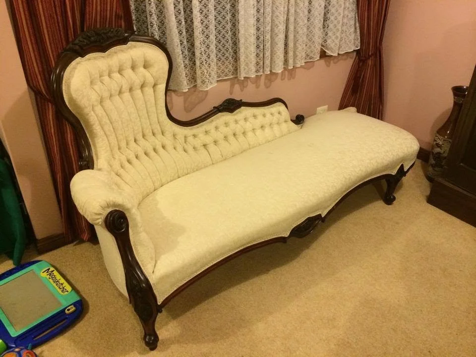 An antique Victorian-style cream-colored upholstered chaise lounge with dark wooden trim and carved legs, positioned in a room with patterned curtains and carpet.