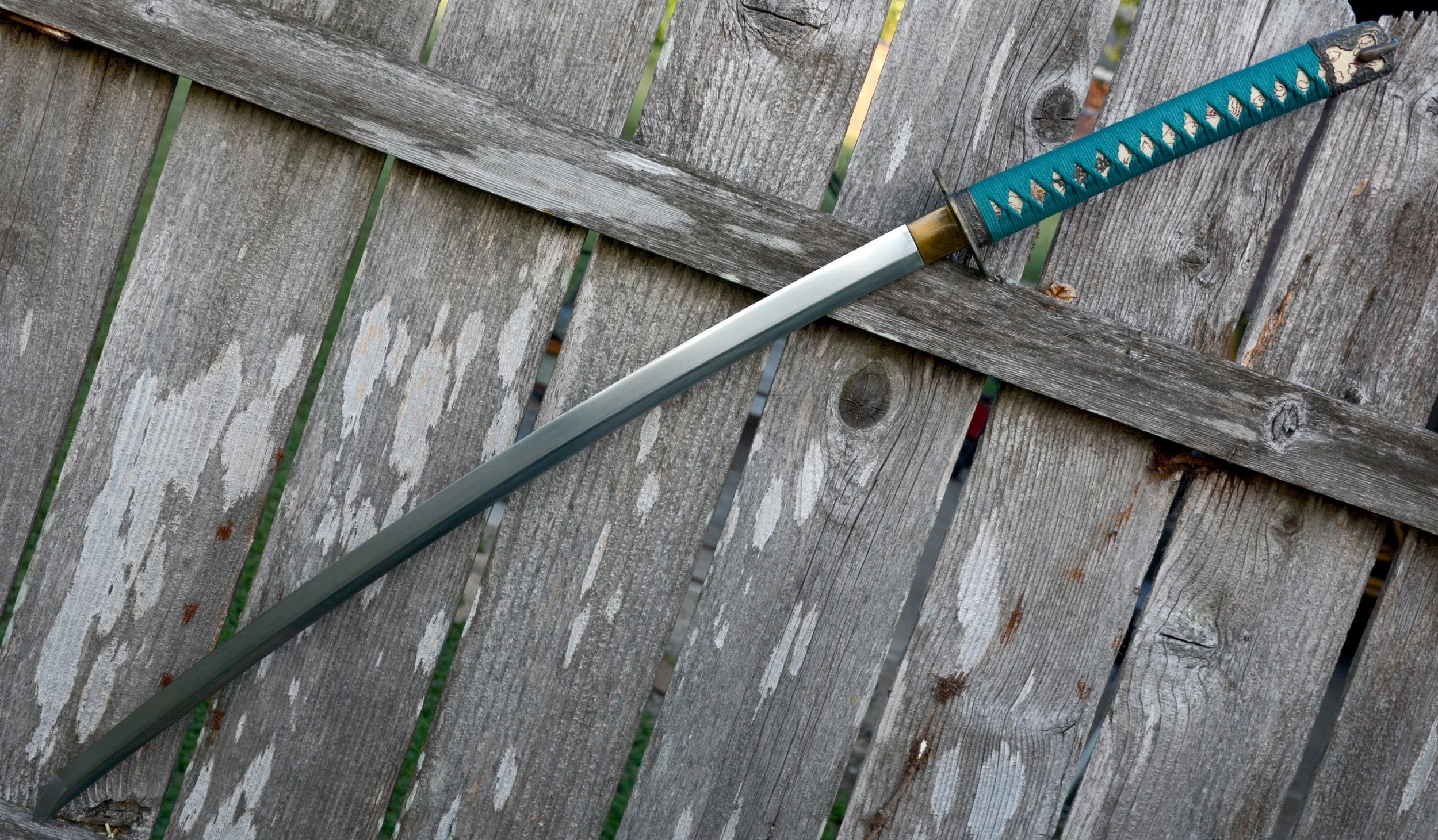 A sword with a teal handle and a metallic blade lies diagonally on a weathered wooden outdoor surface.