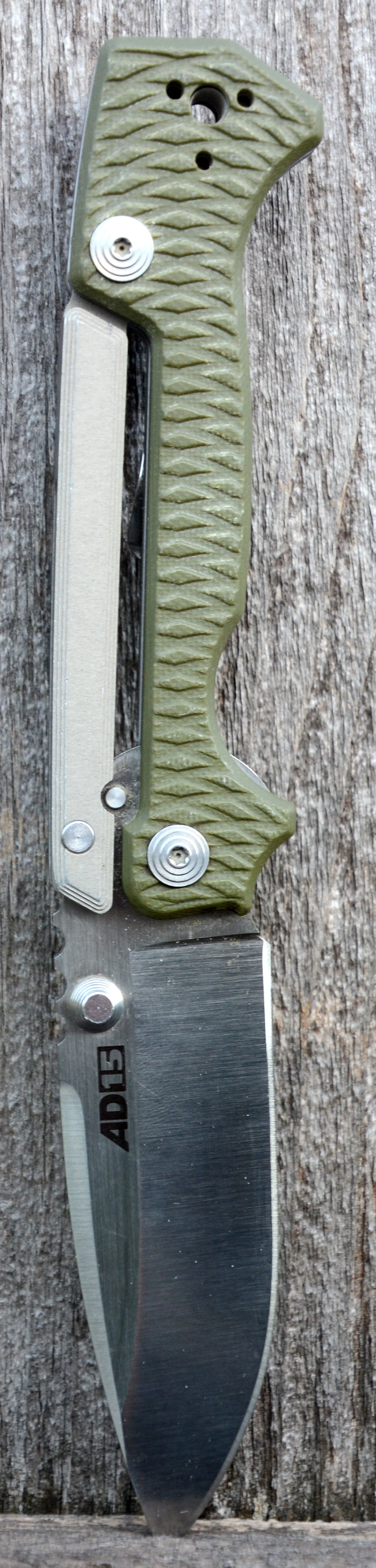 A large fixed-blade hunting or survival knife with a textured green handle, multiple screws, and a black and silver blade with the brand 'AD15' engraved, resting on a weathered wooden surface.