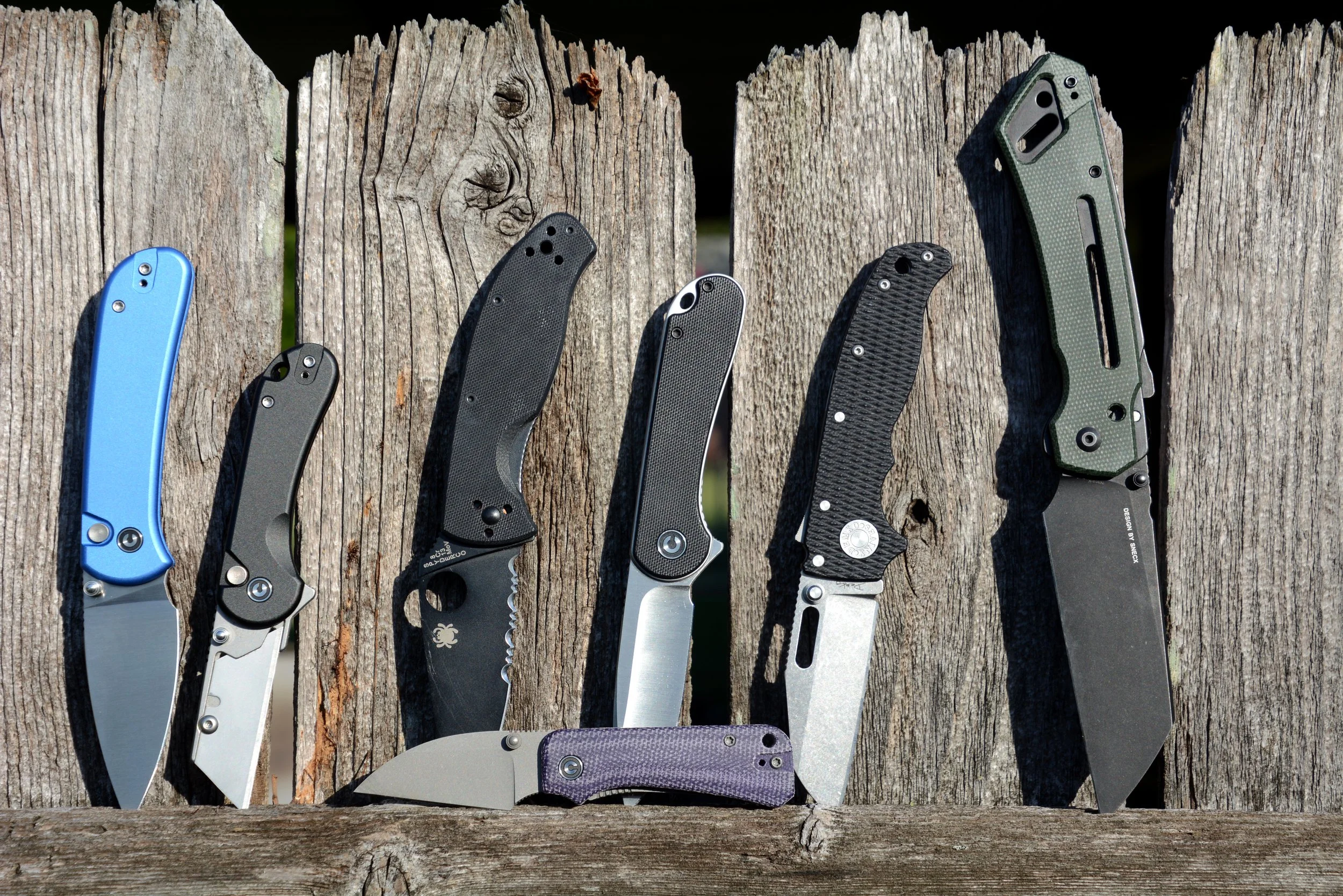 Various folding knives with different handle designs and blade styles are displayed in a row on an aged wooden surface outdoor setting.